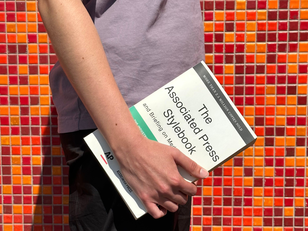 Content strategist Leen holds a printed copy of the AP Stylebook at their side in front of Emspace + Lovgren’s office building, which features a mid-century red, orange and yellow tiled wall.